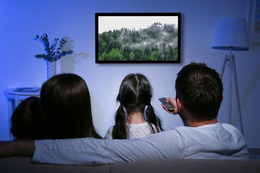 Family of four watching television at home. Back of their head, with dark brown hair shown and TV with trees on display.