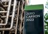 A black sign says 'Zero Carbon 2023' in front of piping 
